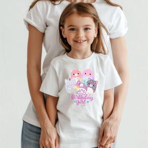 May include: A white t-shirt featuring a colorful graphic of cartoon animals and the text "Birthday girl". The design includes a cat, a pink bird, and other creatures. The shirt is worn by a young girl.