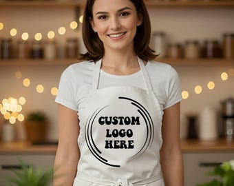 May include: A white apron with the text "CUSTOM LOGO HERE" in a black circular design. The apron has a tie at the waist and a neck strap. The person in the image is wearing a white t-shirt.