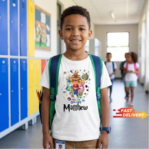 May include: A white short-sleeved t-shirt featuring a colorful graphic print of Super Mario Galaxy characters including Mario, Peach, and Bowser, with the text "The Super Mario Galaxy" and the name "Matthew" printed in a black stylized font.