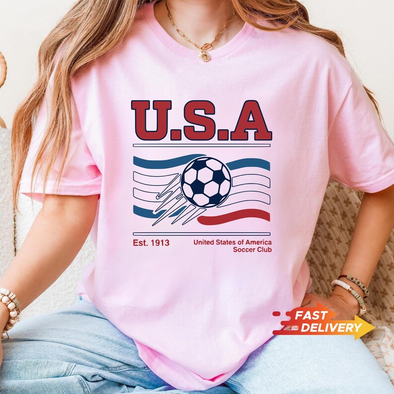 May include: Pink t-shirt with a graphic featuring the text "U.S.A" in red and blue, a soccer ball, and the text "Est. 1913 United States of America Soccer Club". The shirt has short sleeves.