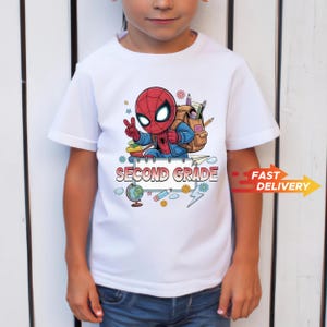 May include: A white short-sleeved t-shirt featuring a graphic of a cartoon superhero wearing a backpack, making a peace sign. Below the character is a notebook graphic with the text SECOND GRADE, surrounded by a globe, pencil, and flowers.