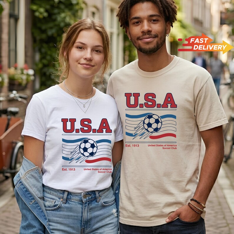 May include: Two people wearing matching t-shirts with a soccer-themed graphic. The shirts feature the text "U.S.A" in red, a soccer ball, and the text "Est. 1913 United States of America Soccer Club". The shirts are white and beige.