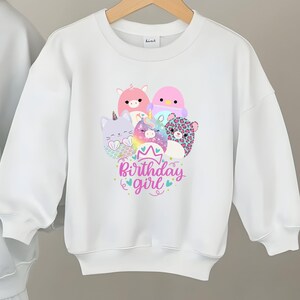 May include: White sweatshirt featuring a colorful graphic of cartoon animals, including a unicorn, cat, and bear. The design includes the text "Birthday girl" in a playful font, with a crown and hearts. The sweatshirt has long sleeves.