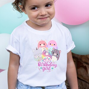 May include: White t-shirt featuring a colorful graphic of cartoon animals, including a cat, unicorn, and leopard, with the text "Birthday girl" in pink and blue. The shirt is worn by a young child.