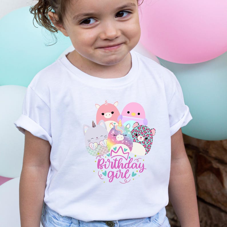 May include: White t-shirt featuring a colorful graphic of cartoon animals, including a cat, unicorn, and leopard, with the text "Birthday girl" in pink and blue. The shirt is worn by a young child.