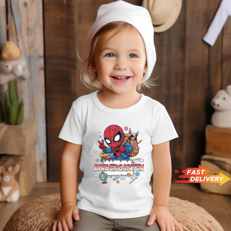 May include: A white short-sleeved t-shirt featuring a graphic of a cartoon Spider-Man character wearing a backpack and making a peace sign. Below the character, the word KINDERGARTEN is printed in bold red letters, surrounded by school supplies.