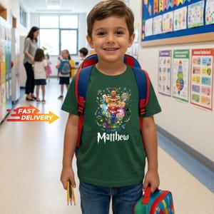 May include: A dark green short-sleeved t-shirt featuring a colorful graphic of Super Mario Galaxy characters including Mario and Bowser, with the name Matthew printed in white text below the design, worn with a red and blue backpack.