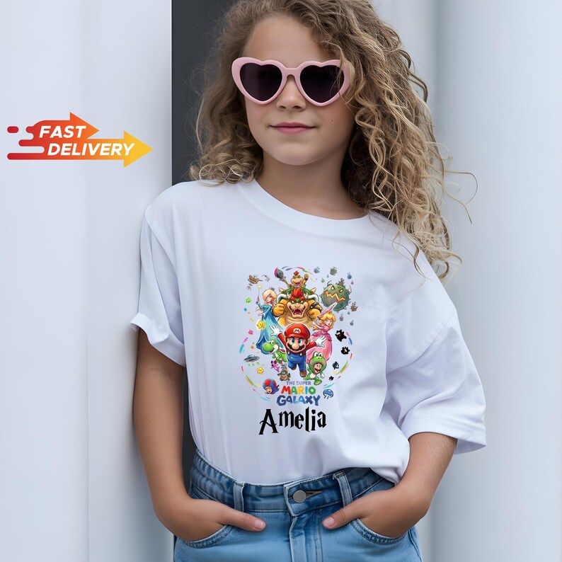 May include: A white short-sleeved t-shirt featuring a colorful Super Mario Galaxy graphic print with various characters and the name Amelia printed in black text below the design. The shirt is paired with blue denim jeans and heart-shaped sunglasses.