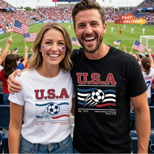 May include: A smiling couple wearing matching t-shirts with a "U.S.A." graphic and a soccer ball design. The shirts are white and black. The background shows a soccer stadium with fans waving American flags.