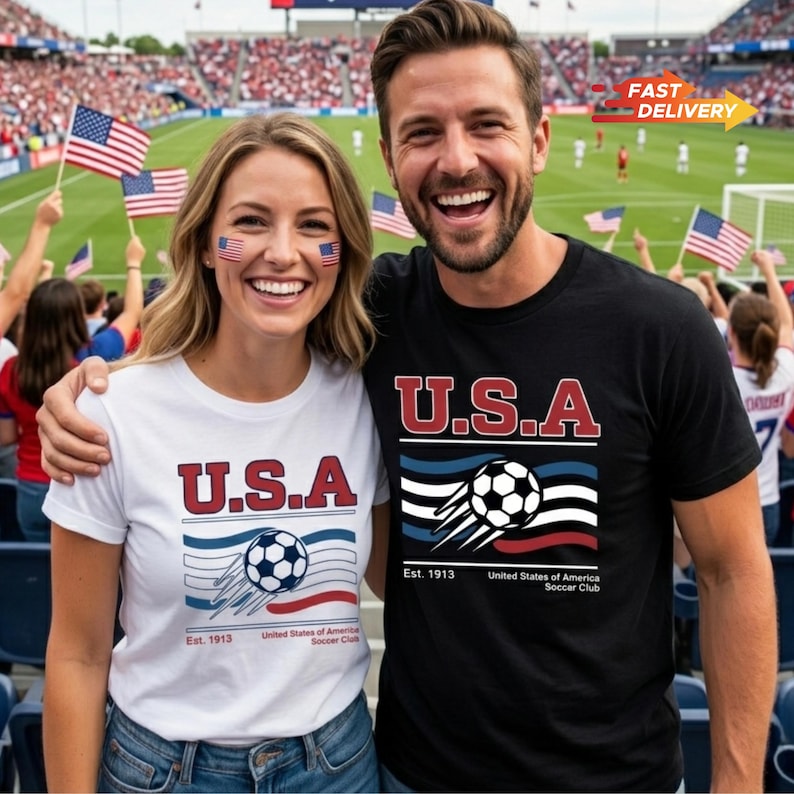 May include: A smiling couple wearing matching t-shirts with a "U.S.A." graphic and a soccer ball design. The shirts are white and black. The background shows a soccer stadium with fans waving American flags.