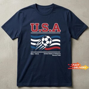 May include: Navy blue t-shirt with a graphic featuring "U.S.A" in red and white lettering above a soccer ball and stylized flag design. The text "Est. 1913" and "United States of America Soccer Club" are below the graphic.