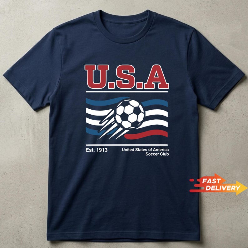 May include: Navy blue t-shirt with a graphic featuring "U.S.A" in red and white lettering above a soccer ball and stylized flag design. The text "Est. 1913" and "United States of America Soccer Club" are below the graphic.