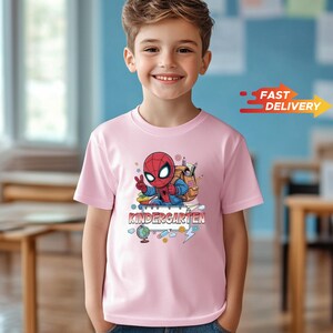 May include: A light pink short-sleeved t-shirt featuring a graphic of a cartoon Spider-Man character wearing a backpack and making a peace sign. Below the character, the word KINDERGARTEN is printed in bold, blocky letters surrounded by school supplies.