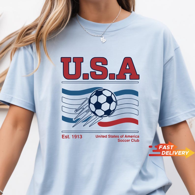 May include: Light blue t-shirt with a graphic featuring "U.S.A" in red block letters above a soccer ball and stripes in red and blue. The text "Est. 1913 United States of America Soccer Club" is below the graphic.