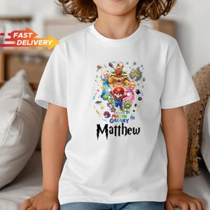 May include: A white short-sleeved t-shirt featuring a colorful graphic of Super Mario Galaxy characters including Mario, Bowser, and Princess Peach. Below the characters, the text reads The Super Mario Galaxy and the name Matthew in black.