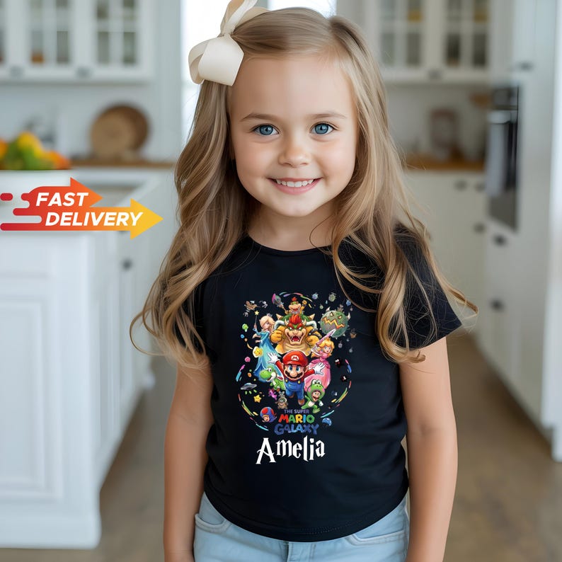 May include: A black short-sleeved t-shirt featuring a colorful graphic print of Super Mario Galaxy characters including Mario, Bowser, and Princess Peach, with the name Amelia printed in white text below the central character illustration.