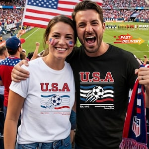 May include: A couple wearing U.S.A. soccer t-shirts, with the American flag in the background. The shirts feature a soccer ball graphic with the text "Est. 1913 United States of America Soccer Club". The woman wears a white shirt, and the man wears a black shirt.