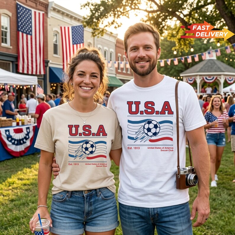 May include: Two people wearing t-shirts with a "U.S.A." soccer design, standing outdoors. The shirts are beige and white, with a soccer ball graphic and the text "United States of America Soccer Club". American flags are in the background.