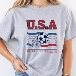 May include: A light gray t-shirt featuring a graphic design with the text "U.S.A" in red, a soccer ball, and the text "Est. 1913 United States of America Soccer Club". The design incorporates blue and red elements.