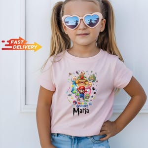 May include: A light pink short-sleeved t-shirt featuring a colorful Super Mario Galaxy graphic print with various characters and the name Maria written in a stylized black font below the design, paired with heart-shaped sunglasses.