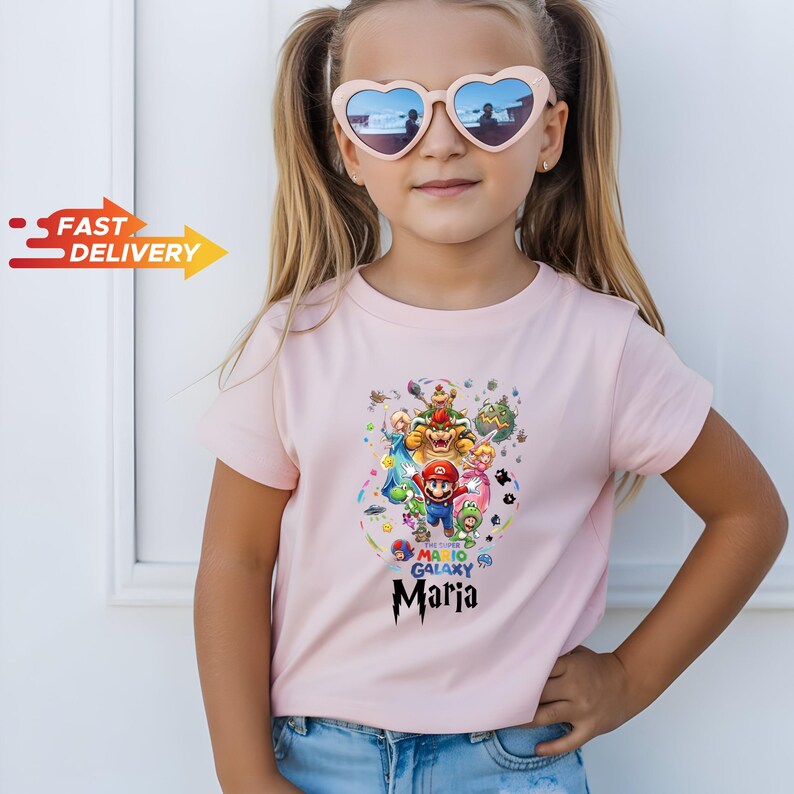 May include: A light pink short-sleeved t-shirt featuring a colorful Super Mario Galaxy graphic print with various characters and the name Maria written in a stylized black font below the design, paired with heart-shaped sunglasses.