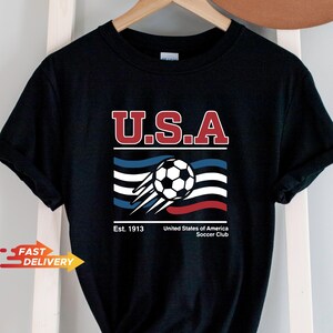 May include: Black t-shirt featuring a soccer ball graphic with the text "U.S.A." in red and white. Below the text is a stylized design with blue, white, and red stripes, and the words "Est. 1913" and "United States of America Soccer Club."