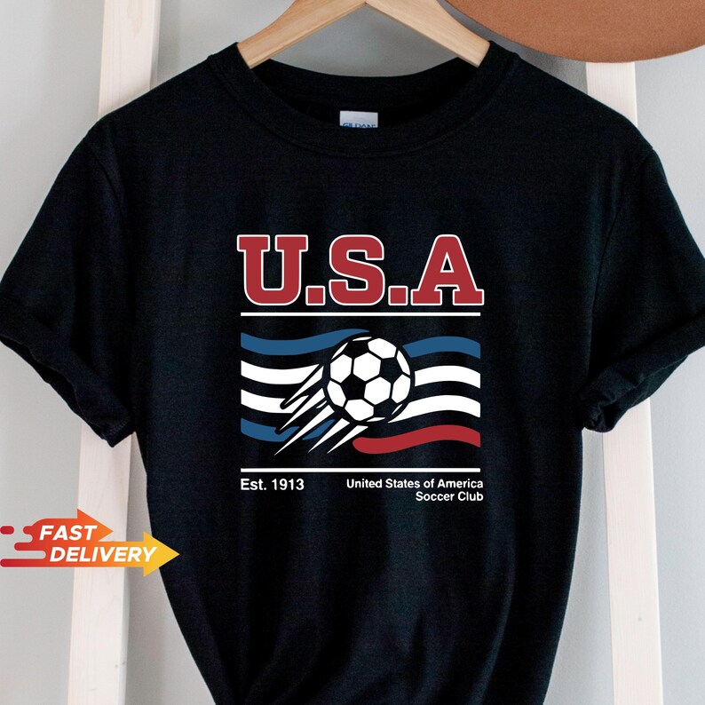May include: Black t-shirt featuring a soccer ball graphic with the text "U.S.A." in red and white. Below the text is a stylized design with blue, white, and red stripes, and the words "Est. 1913" and "United States of America Soccer Club."