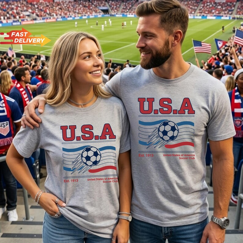 May include: Two people wearing gray t-shirts with the text "U.S.A" in red and blue, along with a soccer ball graphic. The shirts also feature the text "Est. 1913" and "United States of America Soccer Club". They are at a soccer game.