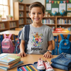 May include: A grey short-sleeved t-shirt featuring a colorful graphic print of Mario, Bowser, Peach, and other characters from the Mario Galaxy series, with the name Matthew printed in a bold black font below the central character illustration.