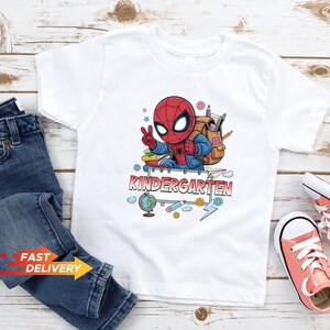 May include: A white short-sleeved t-shirt featuring a graphic of a cartoon Spider-Man character wearing a backpack, making a peace sign, and standing behind a sign that reads KINDERGARTEN, surrounded by school supplies like a globe and pencils.