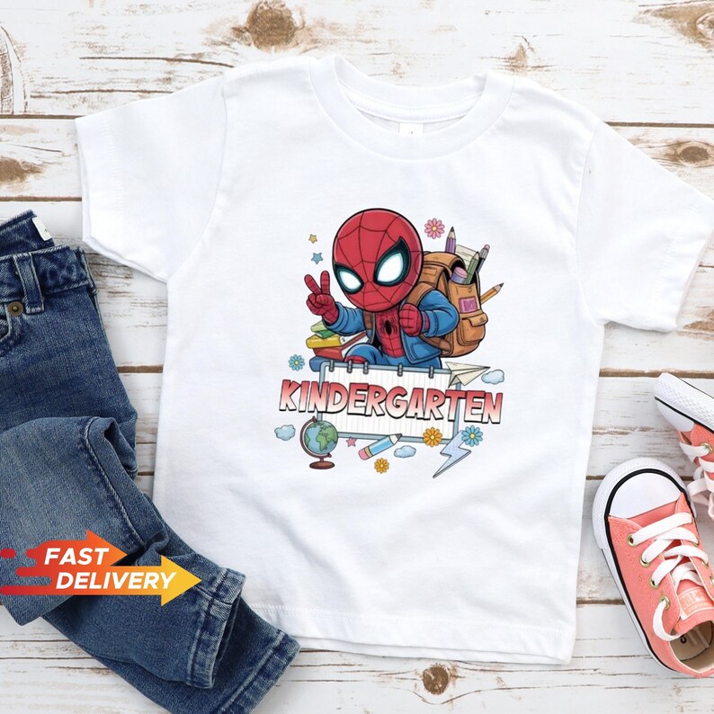 May include: A white short-sleeved t-shirt featuring a graphic of a cartoon Spider-Man character wearing a backpack, making a peace sign, and standing behind a sign that reads KINDERGARTEN, surrounded by school supplies like a globe and pencils.