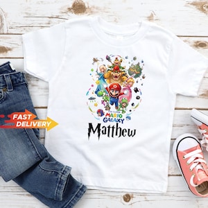May include: A white short-sleeved t-shirt featuring a colorful graphic print of Super Mario Galaxy characters including Mario, Bowser, Princess Peach, and Yoshi. The text reads The Super Mario Galaxy and the name Matthew in a black stylized font.