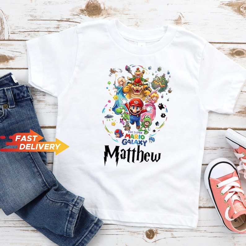 May include: A white short-sleeved t-shirt featuring a colorful graphic print of Super Mario Galaxy characters including Mario, Bowser, Princess Peach, and Yoshi. The text reads The Super Mario Galaxy and the name Matthew in a black stylized font.