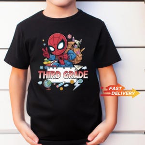 May include: A black short-sleeve t-shirt featuring a graphic of a cartoon superhero wearing a backpack and making a peace sign. Below the character is a notebook page with the text "THIRD GRADE" surrounded by school supplies like a globe and pencil.