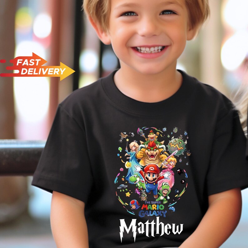 May include: A black short-sleeved t-shirt featuring a colorful graphic print of Super Mario Galaxy characters including Mario, Bowser, and Princess Peach. The text "The Super Mario Galaxy" is printed above the name "Matthew" in white lettering.