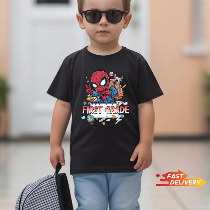 May include: A black short-sleeved t-shirt featuring a graphic of a cartoon superhero character wearing a backpack, with school supplies and the text FIRST GRADE printed in white and red letters on the front of the garment.