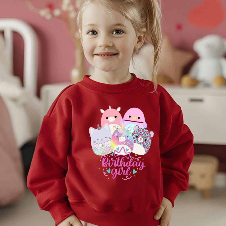 May include: A red sweatshirt featuring a graphic of several cartoon characters and the text "Birthday girl". The design includes a unicorn, cat, and other colorful creatures. The sweatshirt is worn by a young girl.