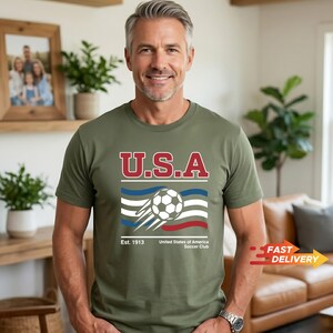 May include: Olive green t-shirt with the text "U.S.A" in red and white. Below the text is a soccer ball graphic with blue and red stripes. The shirt also includes the text "Est. 1913 United States of America Soccer Club."
