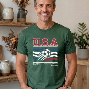May include: Green t-shirt with a graphic featuring the text "U.S.A" in red and white, a soccer ball, and the text "United States of America Soccer Club". The shirt has short sleeves.