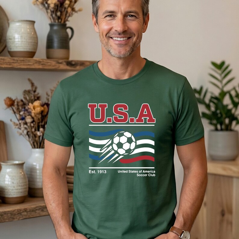May include: Green t-shirt with a graphic featuring the text "U.S.A" in red and white, a soccer ball, and the text "United States of America Soccer Club". The shirt has short sleeves.