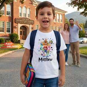 May include: A white short-sleeved t-shirt featuring a colorful graphic of Mario and various characters from the Super Mario Galaxy series, with the name Matthew printed in a black stylized font below the characters, worn with a dark blue backpack.