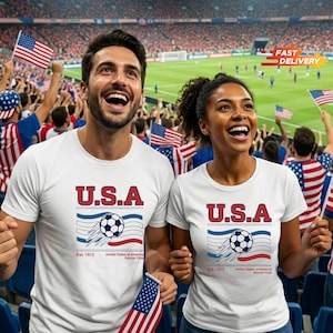 May include: Two people wearing white t-shirts with a "U.S.A." soccer design, waving American flags in a stadium. The shirts feature a soccer ball graphic, blue and red stripes, and the text "Est. 1913 United States of America Soccer Club."