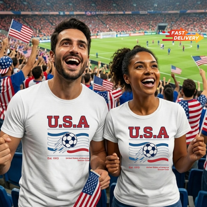May include: Two people wearing white t-shirts with a "U.S.A." soccer design, waving American flags in a stadium. The shirts feature a soccer ball graphic, blue and red stripes, and the text "Est. 1913 United States of America Soccer Club."