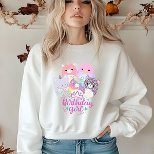 May include: White sweatshirt featuring a colorful graphic of cartoon animals, including a unicorn, cat, and leopard. The text "Birthday girl" is written in pink and blue script. The sweatshirt is a casual style.