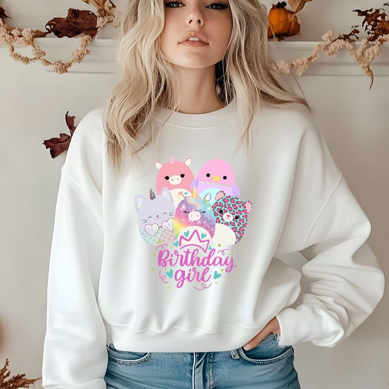 May include: White sweatshirt featuring a colorful graphic of cartoon animals, including a unicorn, cat, and leopard. The text "Birthday girl" is written in pink and blue script. The sweatshirt is a casual style.