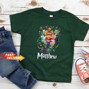 May include: A dark green short-sleeve t-shirt featuring a colorful graphic print of Super Mario Galaxy characters including Mario, Bowser, and Princess Peach. The text "The Super Mario Galaxy" and the name "Matthew" are printed in white below the graphic.