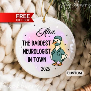 Puede incluir: Adorno navide&ntilde;o de cer&aacute;mica blanca con una cuerda dorada para colgar. El adorno presenta el texto "Alex THE BADDEST NEUROLOGIST IN TOWN 2025" y un ganso de dibujos animados con uniforme y mascarilla. Una pancarta roja dice "FREE GIFT BOX".