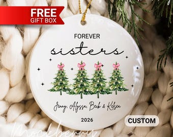 May include: A white ceramic Christmas ornament with the words "FOREVER sisters" in black script. Four green Christmas trees with pink bows are below the text. The names Jenny, Alyssa, Beck & Kelsea and the year 2026 are printed below the trees. A gold string is attached.