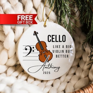 May include: A white ceramic ornament with a cello illustration and the text "CELLO LIKE A BIG VIOLIN BUT BETTER". The ornament also includes the name "Anthony" and the year "2025". A gold string is attached for hanging.