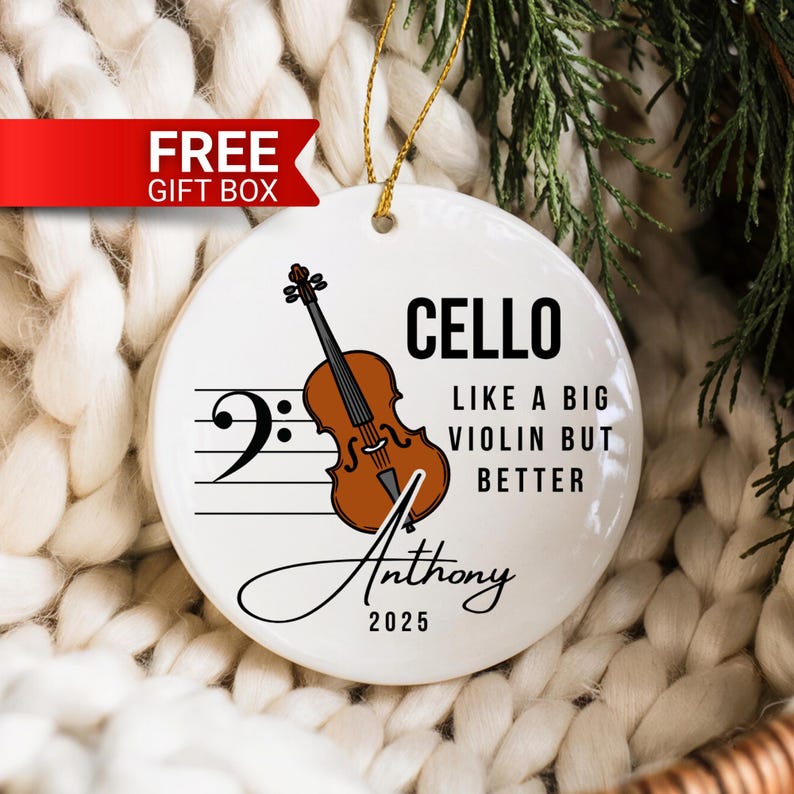 May include: A white ceramic ornament with a cello illustration and the text "CELLO LIKE A BIG VIOLIN BUT BETTER". The ornament also includes the name "Anthony" and the year "2025". A gold string is attached for hanging.
